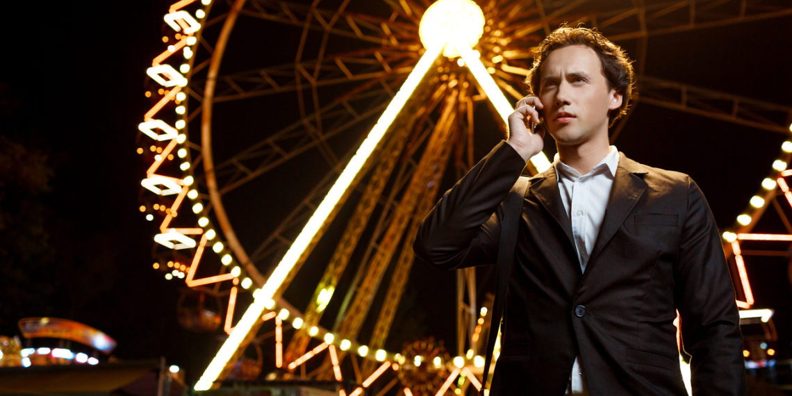 Portrait of young successful businessman in suit speaking on phone over night amusement park.