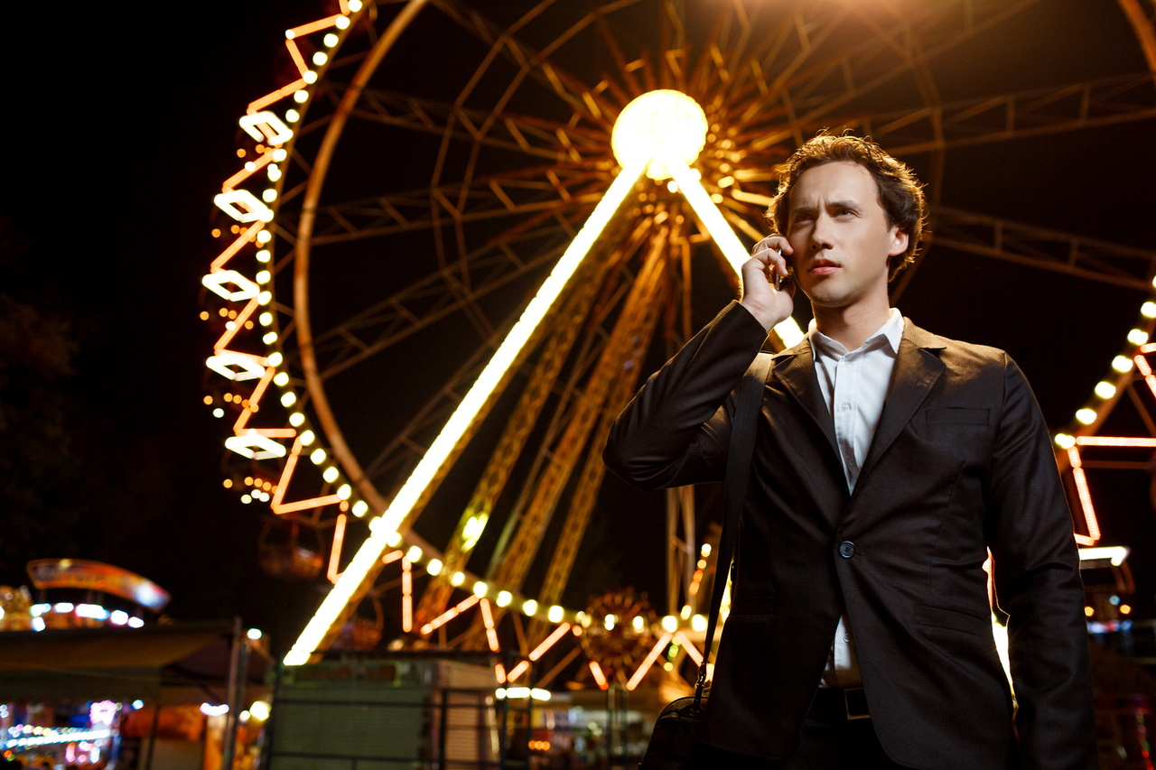 Portrait of young successful businessman in suit speaking on phone over night amusement park.